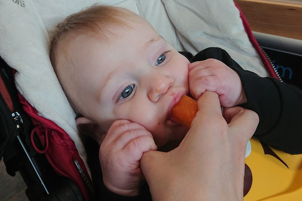 A baby eating watermelon. His mum is holding the watermelon and he is holding her fingers to guide it.