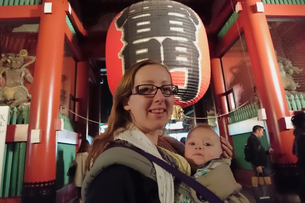 A picture of a mother carrying her baby in a carrier. Both looking at the camera while stood in front of a giant red lantern