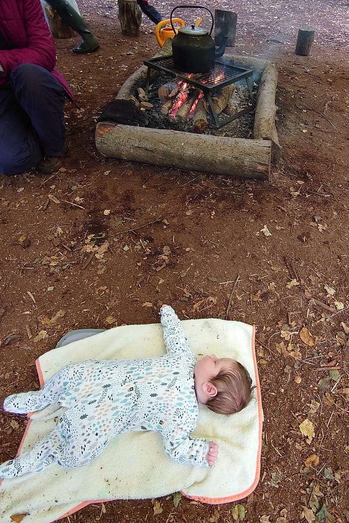 A little baby lying on a blanket on the forest floor, watching a fire